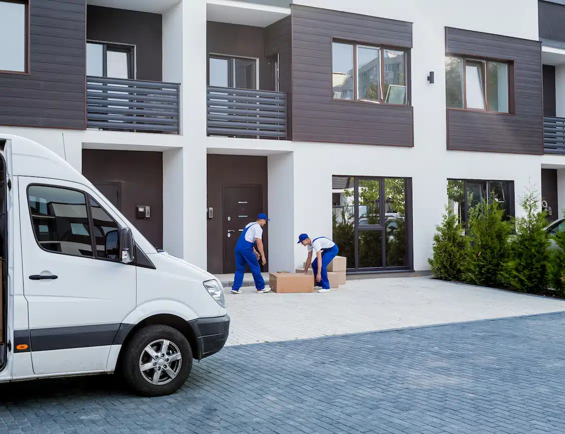On the Hook Agency website: Two skilled movers in blue uniforms from a trusted moving company efficiently unload a large cardboard box from a white van positioned outside a contemporary apartment complex. The stylish building showcases sleek white walls, dark balconies, and neatly arranged shrubs enhancing the entrance.