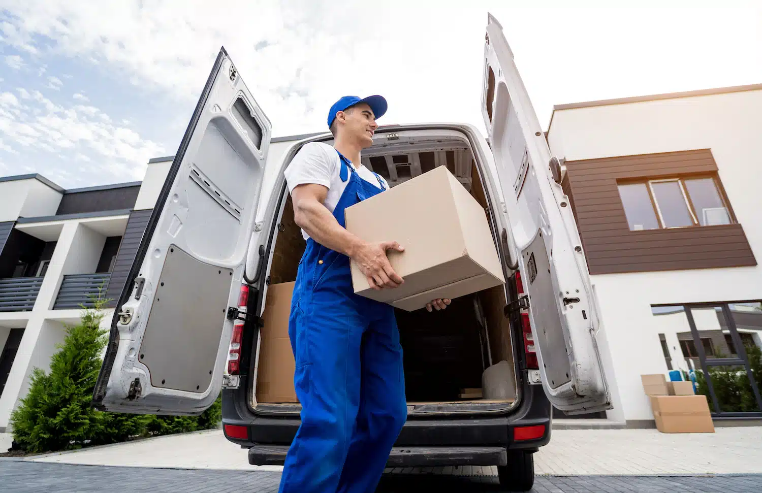 A delivery professional, clad in blue coveralls and a cap, efficiently unloads a company-branded cardboard box from an open van amidst a residential neighborhood.