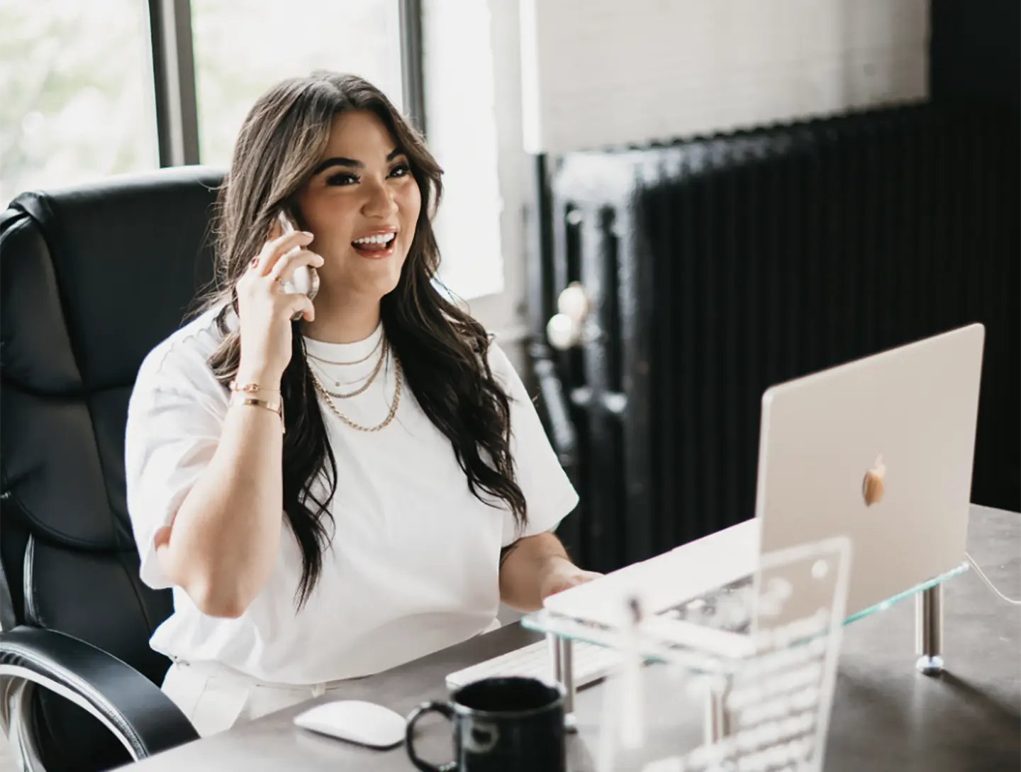 A woman with long dark hair, wearing a white shirt and layered necklaces, sits at a desk engaged in a phone call. She smiles as she works, with a laptop open before her and a mug and mouse nearby, embodying her daily routine at Hook Agency.