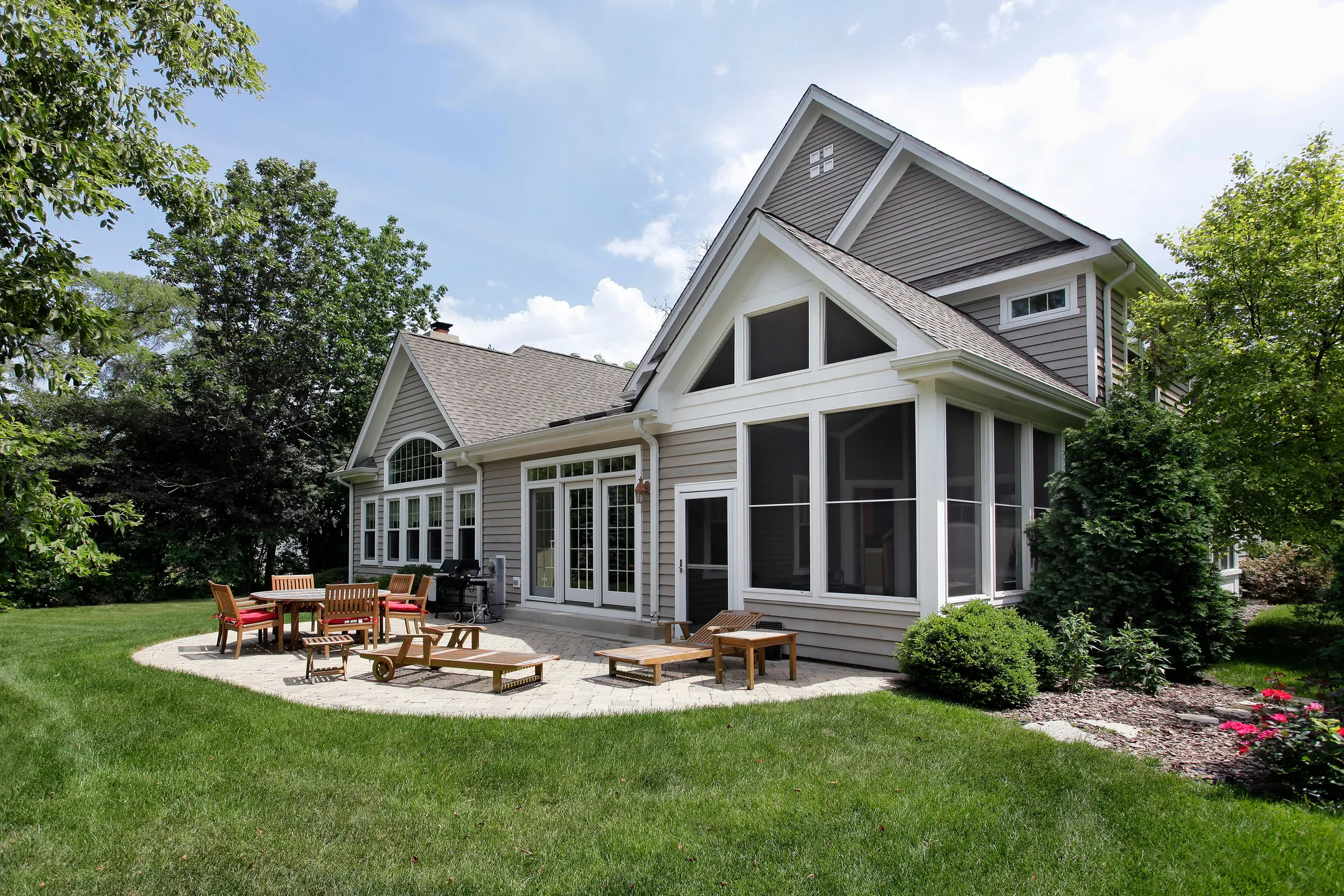 A modern suburban house with large windows, a screened porch, and a gabled roof. The backyard features a stone patio, crafted by expert patio contractors, with wooden chairs and tables, surrounded by a well-maintained lawn and trees under a partly cloudy sky.