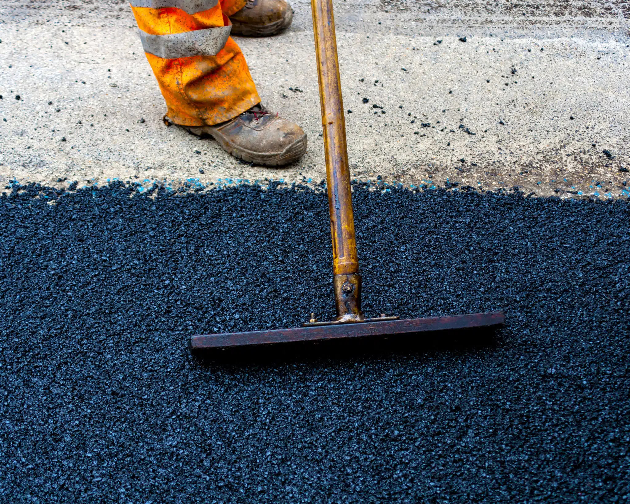 On Hook Agency's website, an image captures a paving worker in orange reflective pants and brown boots expertly smoothing fresh black asphalt with a wooden-handled tool. Above, a completed section of road showcases its flawless texture, effectively illustrating the superior quality of seamless roadwork.