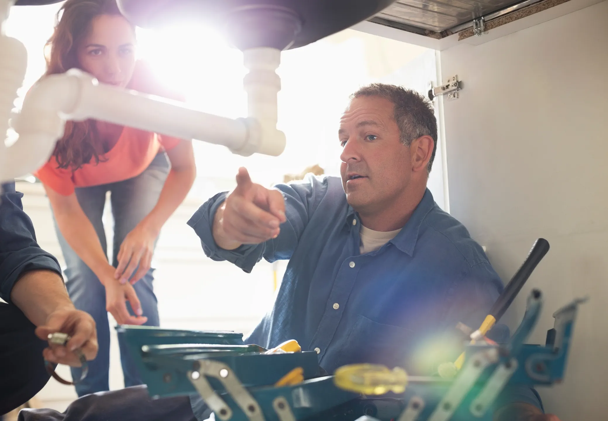 A man under a kitchen sink demonstrates a plumbing task to an attentive woman beside him. Tools are scattered around as sunlight streams through the window, creating a warm glow.