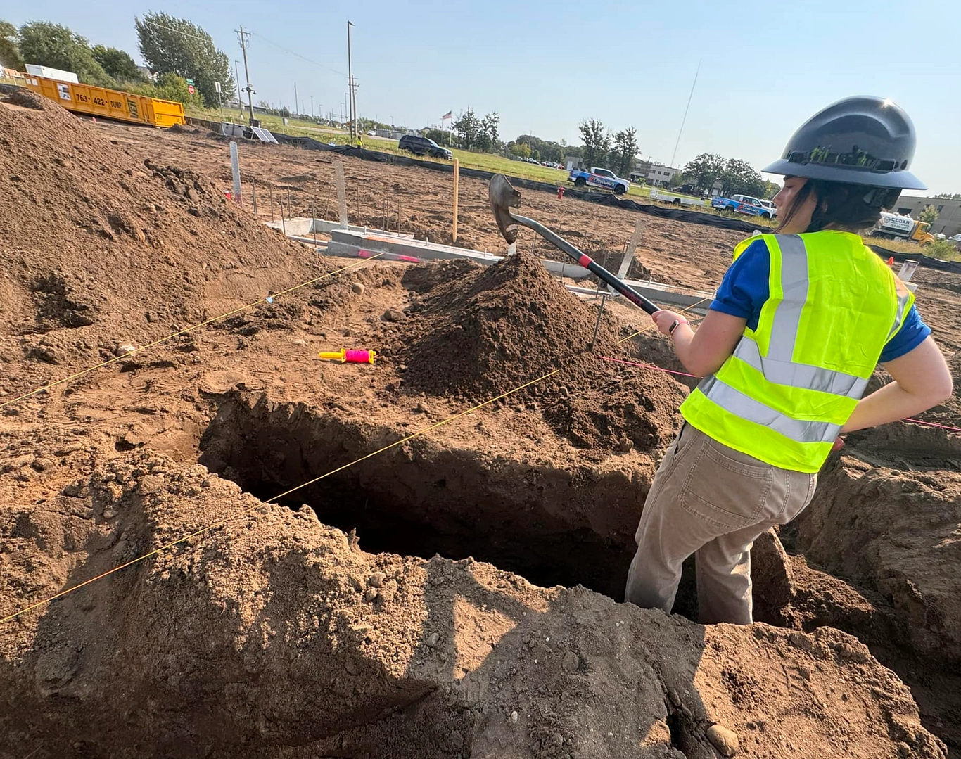 A construction worker in a safety vest and helmet shovels dirt at an active excavation site, preparing the ground for plumbing installation. Large soil piles, measuring tapes, and work vehicles surround the area—perfect imagery to feature on your plumbing website to convey professionalism and expertise.