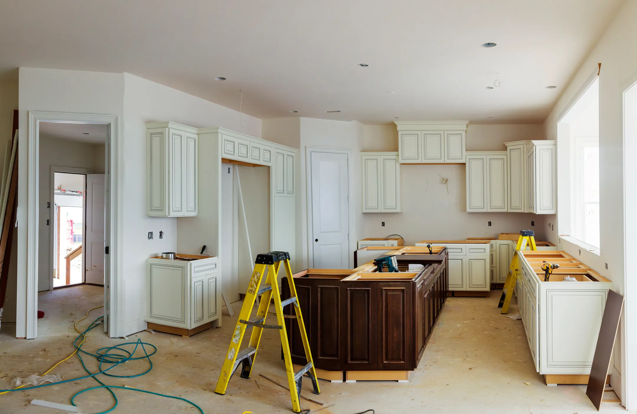 A kitchen renovation features white cabinets and a central dark wood island, accompanied by two yellow ladders. Amidst scattered tools and materials, visible wires and light fixtures indicate ongoing work. Large windows illuminate the space with natural light, symbolizing an effective digital marketing strategy in design execution.