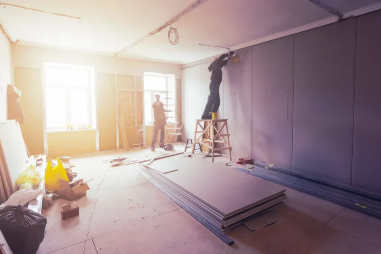 Amidst a dynamic remodeling project, two individuals diligently transform a sunlit room. One installs fixtures from a ladder near the ceiling, while the other systematically organizes materials on the floor. Drywall sheets and construction tools are strategically positioned as part of this ongoing renovation.