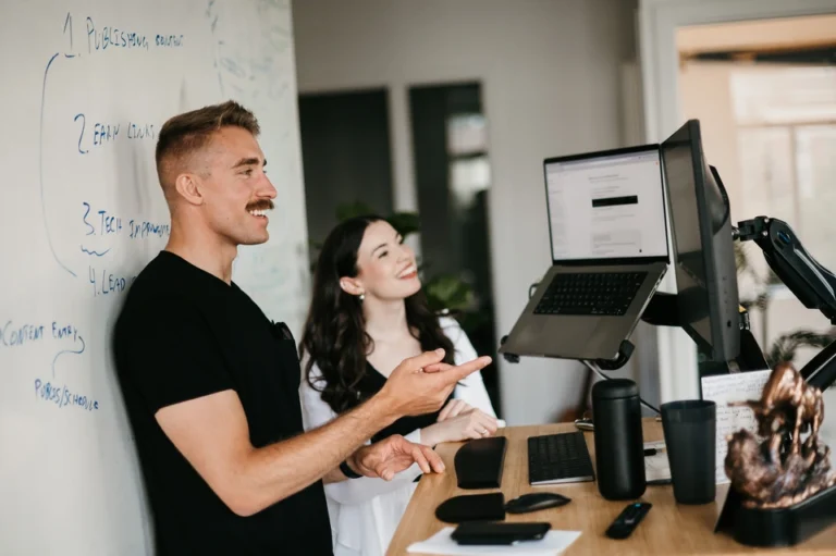 On Hook Agency's website, a man and woman collaborate at a desk, focused intently on Minnesota SEO strategies. The man gestures towards the computer screen, while the woman smiles, reflecting their productive exchange. A whiteboard filled with diagrams enhances the dynamic environment of their discussion.