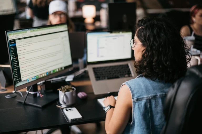 A woman with curly hair and glasses diligently works at her desk, efficiently managing Fire Restoration data on a large monitor and laptop.