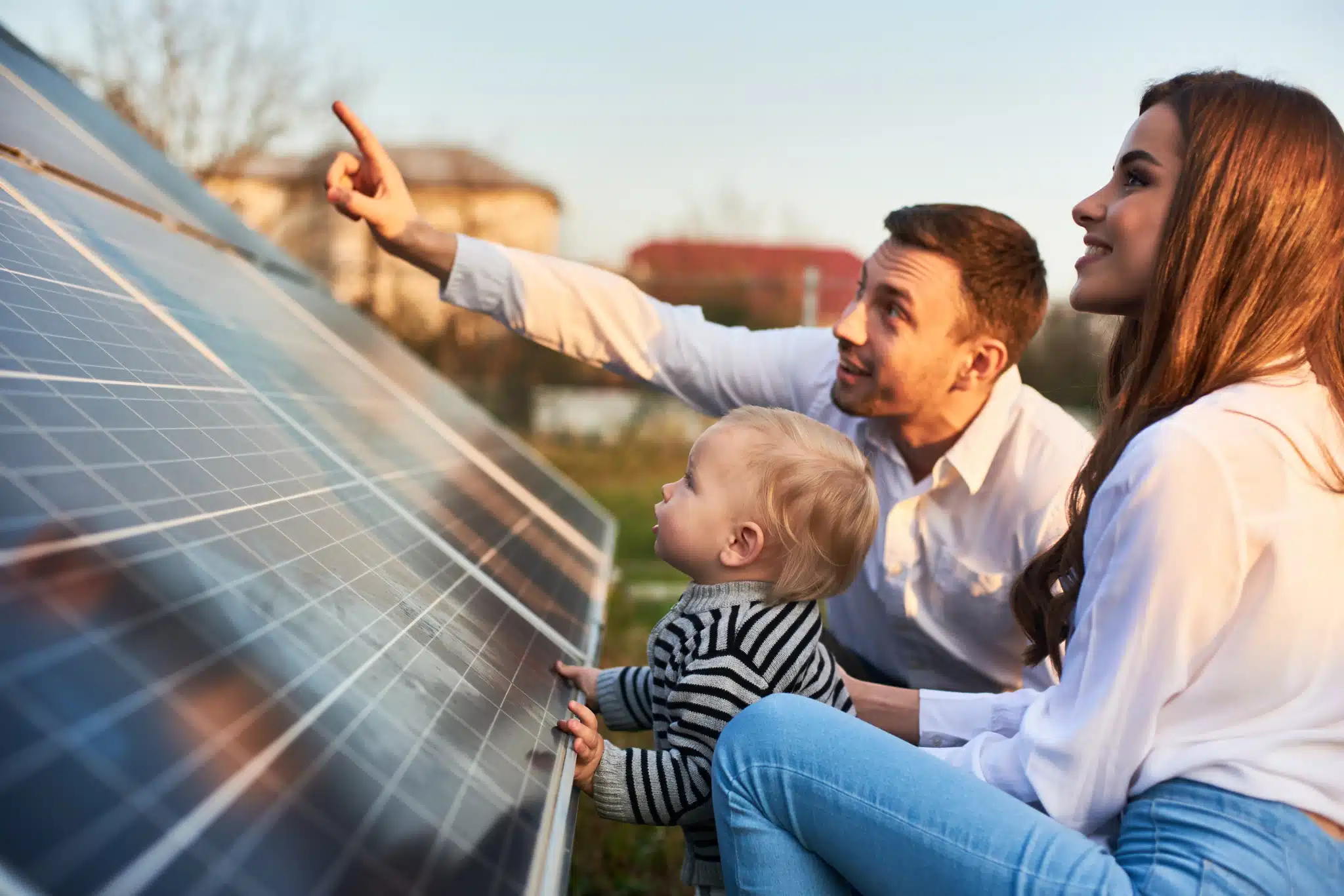 A family—man, woman, and toddler—relax beside a solar panel under a clear sky. The man gestures into the distance, embodying harmony and innovation. This scene is ideal for marketing campaigns centered on sustainable living.