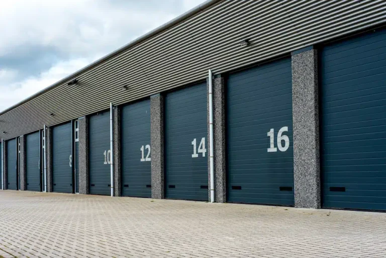 A series of industrial garage doors marked 10, 12, 14, and 16 line a storage warehouse complex. The building features a corrugated metal roof and is set against a paved foreground beneath an overcast sky.