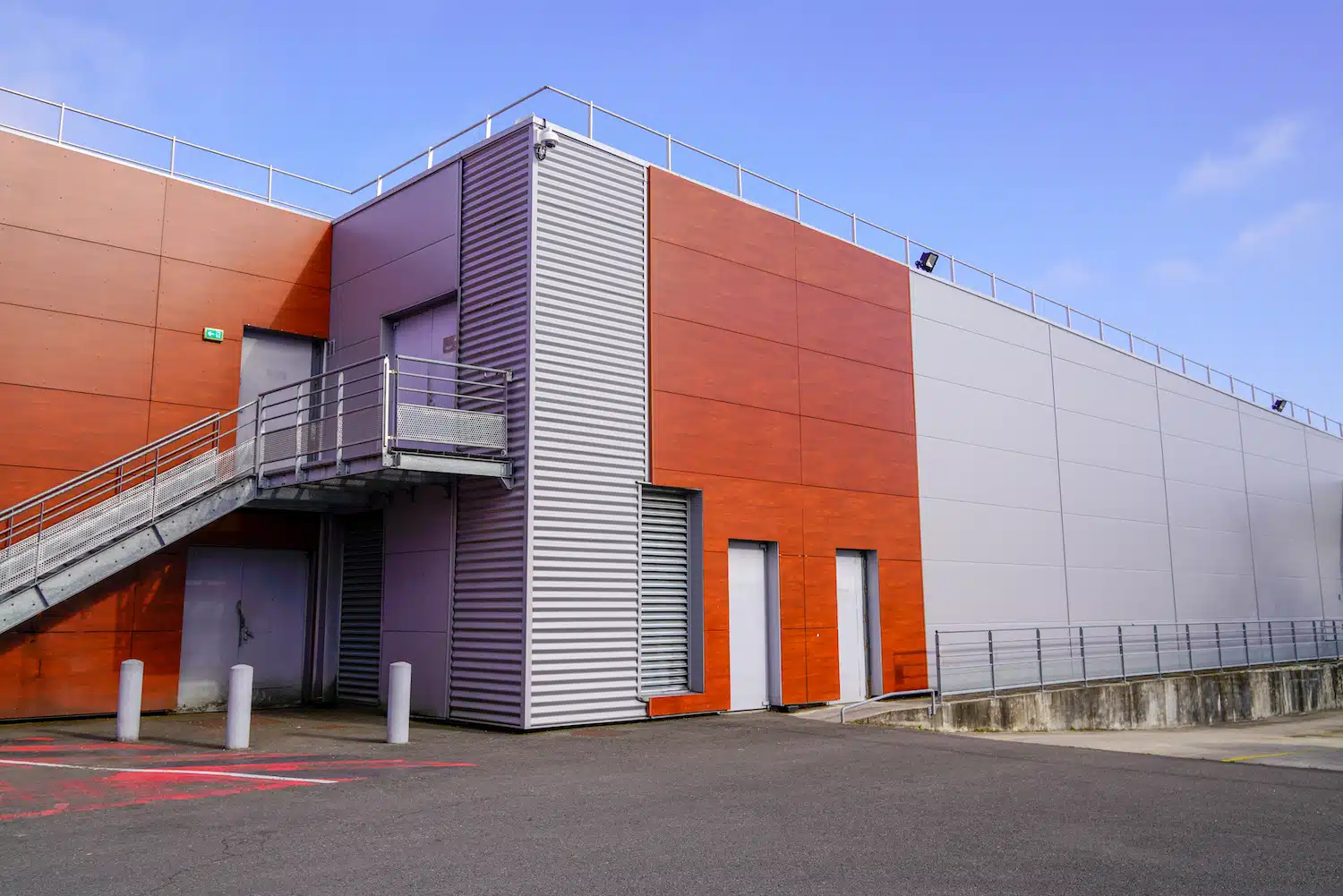Discover a modern industrial facility featuring striking red and gray panels, complete with a metal staircase and designated storage zones. Several entry points to various businesses open onto a paved area, all framed by a pristine blue sky.