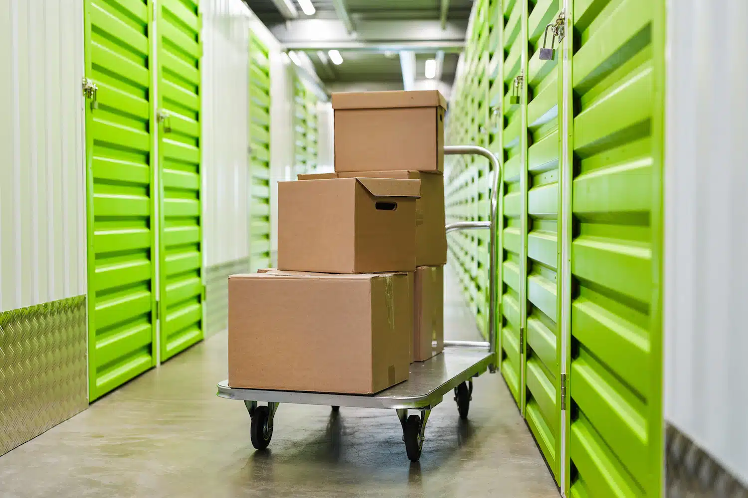 A cart loaded with stacked cardboard boxes stands ready for transport in a well-lit storage facility, flanked by green doors on either side.