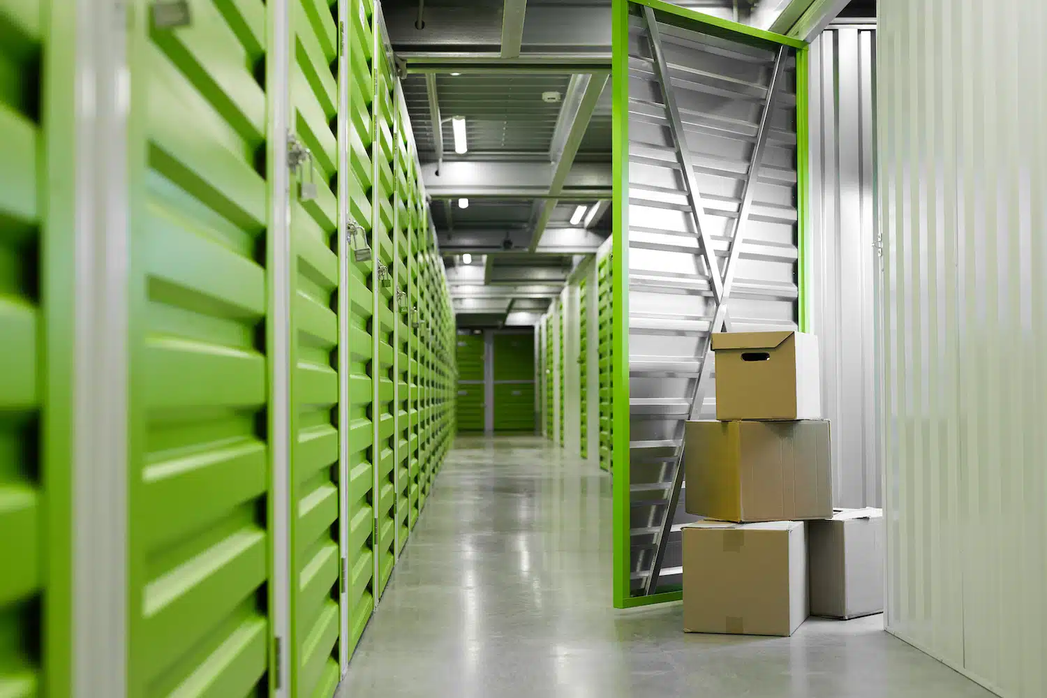 Experience the excellence of a premier storage company: vibrant green doors line the hallways of our facility, embodying quality and security. Open doors unveil spacious, adaptable storage solutions. Nearby, three stacked cardboard boxes rest on polished concrete floors under bright industrial lighting, ready for your storage needs.