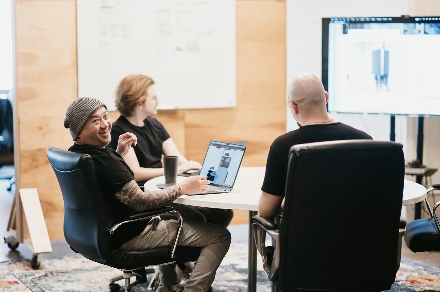 Three professionals gather in a well-equipped meeting room around a round table. One of them smiles as he holds his laptop, while a whiteboard and large monitor provide the perfect backdrop for their collaborative discussion.