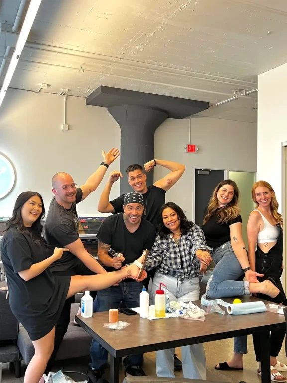 Six individuals gather, envisioning futures in tattoo artistry, confidently showcasing temporary tattoos on their arms. The room, equipped with tattoo supplies, includes a cozy couch and benefits from natural light streaming through a large window, all under overhead lighting.