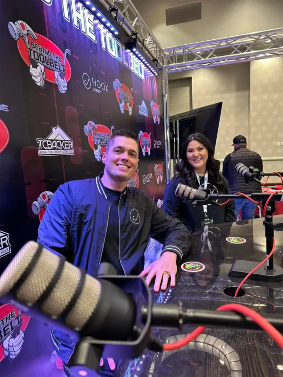 Two individuals sit poised at a table, microphones ready, against a backdrop showcasing logos and the phrase "Behind the Toolbelt." Engaged and prepared, they are set to delve into discussions about their careers, accompanied by promotional materials on the table.