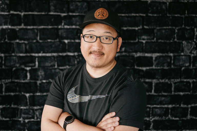 A person in a black Nike shirt and glasses stands confidently against a dark brick wall, wearing a black cap with the brown Tim Brown logo.