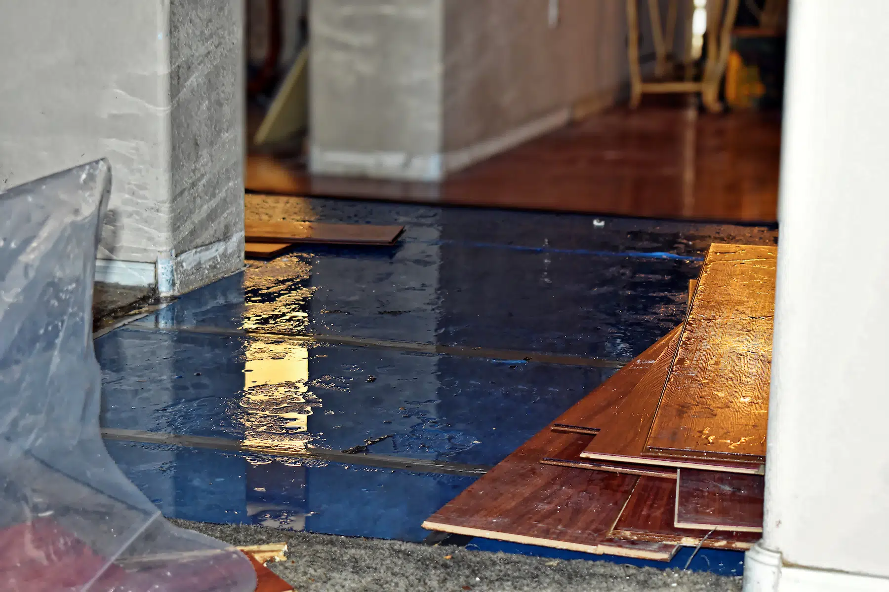 A partially flooded room with water pooling over part of the floor resembles an abstract painting. On one side, stacked hardwood flooring appears like a unique marketing exhibit, while a clear plastic sheet is situated to the left. In the background, an adjoining room features darker flooring.