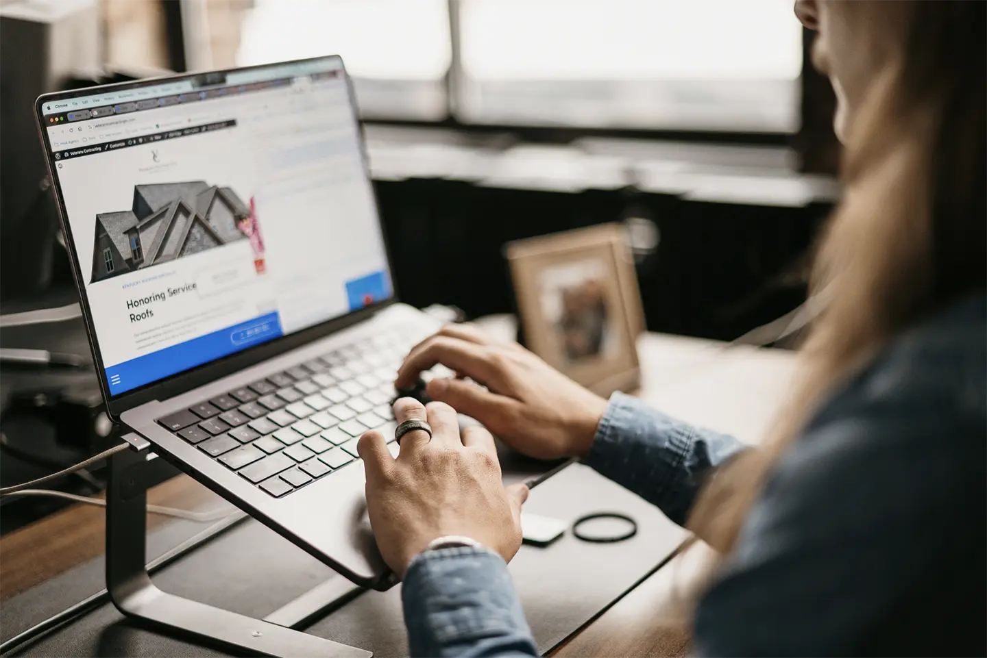 A user explores dream homes on a website displayed on a laptop resting on a desk stand. Beside the laptop, a small photo frame and ring lie quietly, enhancing the peaceful ambiance created by soft natural light streaming through the window.