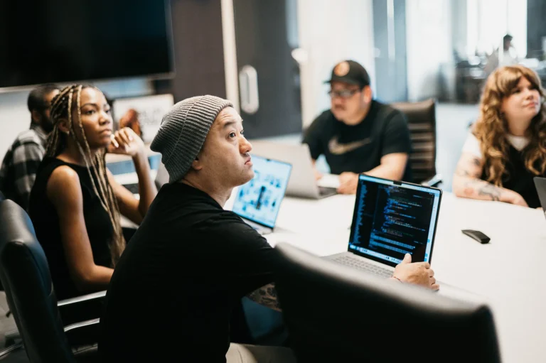 A focused team gathers around a conference table, actively discussing strategies. Open laptops reveal website design code, while a large screen displays SEO keyword optimization tactics, underscoring their commitment to enhancing online visibility.