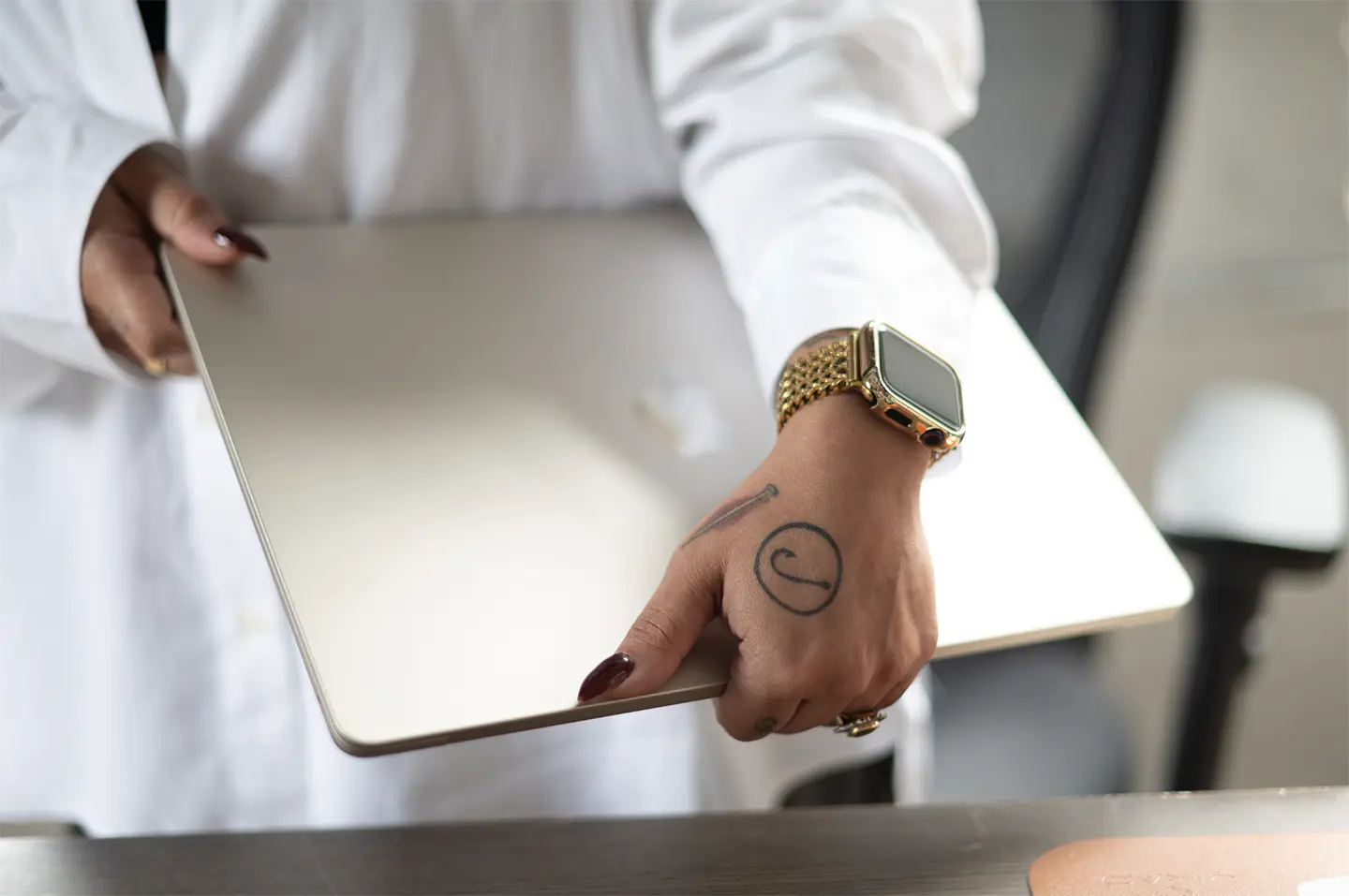 A person with a gold watch and hand tattoo captures the essence of modern work by holding a closed laptop. Dressed in a white long-sleeve shirt, they stand beside a desk featuring a brown item, highlighting the hand and laptop.