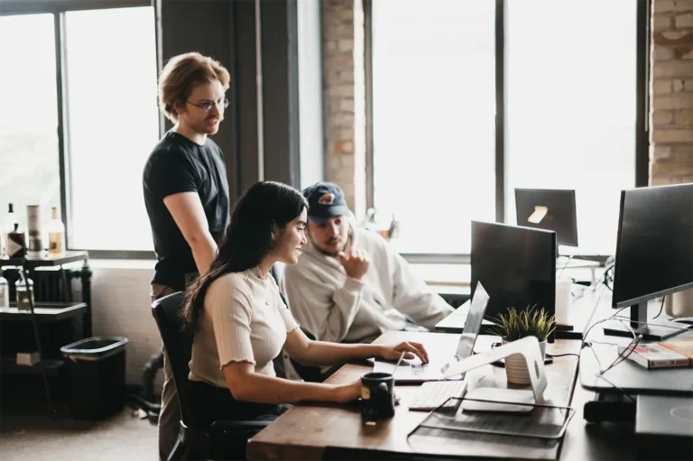 In a well-lit, modern office with large windows at Hook Agency, a seamless collaboration unfolds: a woman focuses on her laptop while two men attentively observe, fostering productivity and innovation.